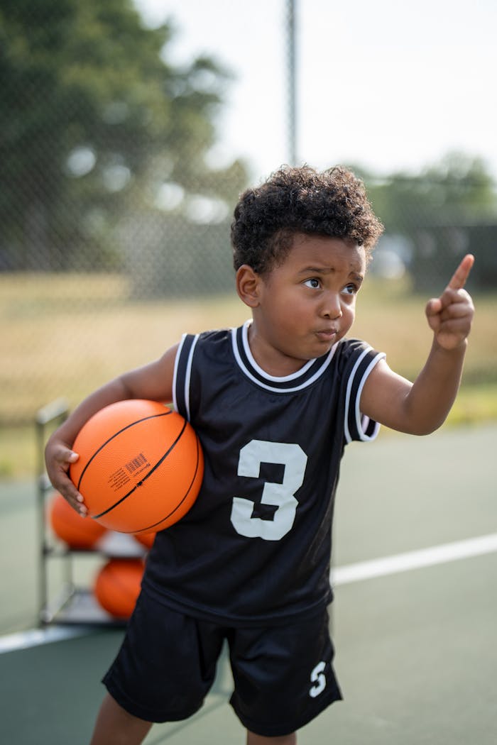 A young boy holding a basketball on an outdoor court, pointing energetically.